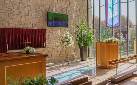 Inside the chapel at Salisbury Crematorium — stone walls, a stained glass window, a wooden lectern and benches, with garden visible through full-height glazing